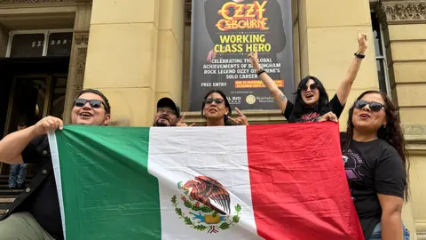 Two men and three women posing with the Mexico flag in front of Birmingham Museum and Art Gallery  and a sign for the Ozzy Osbourne exhibition