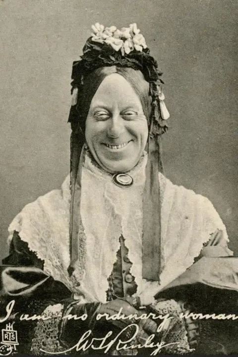 Print Collector/Getty Images A black and white portrait of a Arthur Lucan, dressed as a washerwoman and smiling at the camera. He is wearing a white cloth, draped over both of his shoulder with a circular brooch in the middle. He is also wearing a long fabric hat with flowers at the top.