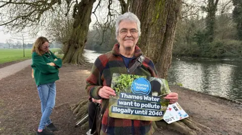 Martin Heath/BBC Keith Dancy with short white hair smiling at the camera and wearing a coat of coloured square patches. He is holding a poster which says "Thames Water wants to pollute illegally until 2040". A person with long brown hair wearing a green sweater and blue trousers is standing cross-legged behind him. They are standing on mud in front of two trees.