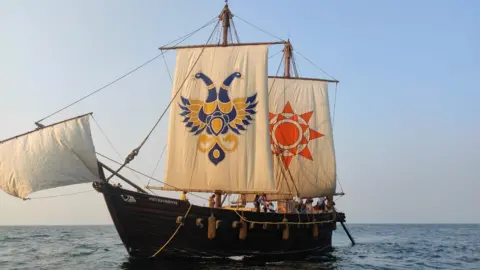 A hand-stitched wooden sailing ship with two large cream-coloured square sails, decorated with motifs is seen mid-sea with crew members on the deck.