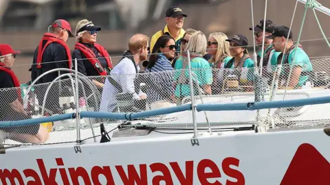 Getty Images Harry and Meghan seen from behind on a large white vessel with 'Making waves' printed on the side in red letters. On the right side sits six people wearing turquoise T-shirts and life jackets, and on the left sits three people in dark clothes and red life jackets. Meghan is wearing a navy and white striped shirt with sunglasses and Harry wear a light-coloured shirt. Both wear dark life jackets.