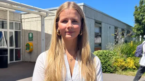 Amelie pictured with long blonde hair looking directly at the camera. She is wearing a white blouse and gold hoop earrings.