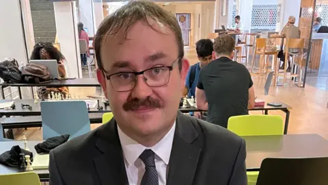 A man with short dark hair and a moustache, wearing glasses and a black suit, pictured from the shoulders up with chessboards behind him and people sat behind him playing chess.