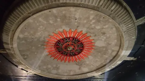 A large dome on the ceiling of The Millennium building. The grey domed is carved and has an orange, red and black feature in the middle of it.