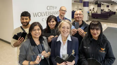 A group of seven people stand in an office holding mobile phones with a City of Wolverhampton Council logo behind them