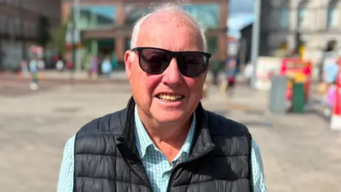 A man wearing sunglasses, gilet and blue shirt