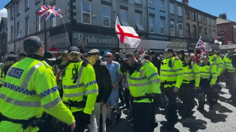 Jonny Humphries/BBC A line of police officers in yellow reflective jackets walk alongside a large group of protesters, some of who are carrying English and union jack flags. Beyond are shops with only the upper floors visible above the crowd. It's sunny, so the officers are casting distinct shadows on the grey tarmac of the road.