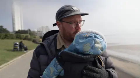 A man in a hat and glasses looks towards the sea. He is holding a child in a carrier