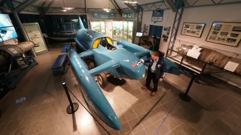 Donald Campbell's restored Bluebird hydroplane - a large jet-powered blue craft - is polished by a museum volunteer as it sits on display at the Ruskin Museum. Other items of Campbell memorabilia decorate the room's walls.