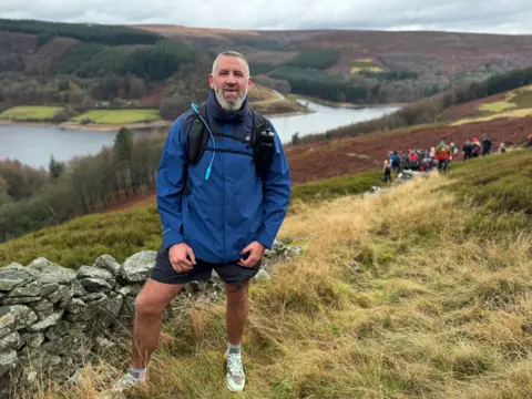 Jason is pictured on the walk with Derwent Reservoir and the Derbyshire countryside behind him 