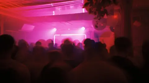 Inside Cobalt during a club night which is filled with people who are slightly blurred and cast in shadow. The lights are pink and there is a light up lit up rectangular structure towards the back of the room. 