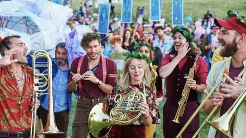 A group of men and women at a festival holding brass instruments. A blonde women is in the middle wearing a flowery top and holding a french horn. Around her, other musicians are holding trombones and trumpets. They are all wearing green leaf head crowns. in the background are other festival-goers on a green landscape