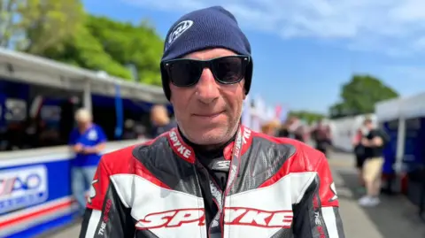 Helge Larson is wearing red, black, and white leathers. He has short stubble and is wearing a blue beanie hat and sunglasses.