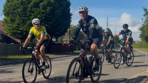BBC/Jim Scott Moody and several other people on bikes cycle along the road. 