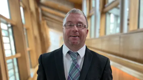 Robert White is in a Scottish Parliament corridor and is smiling broadly at the camera. He has short, grey hair and is wearing a black suit jacket, white shirt and a purple and turquoise tartan tie.