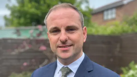 A man with short brown and grey hair, he is smiling at the camera and is wearing a navy suit, blue shirt and grey spotted tie. The background behind him is blurred.