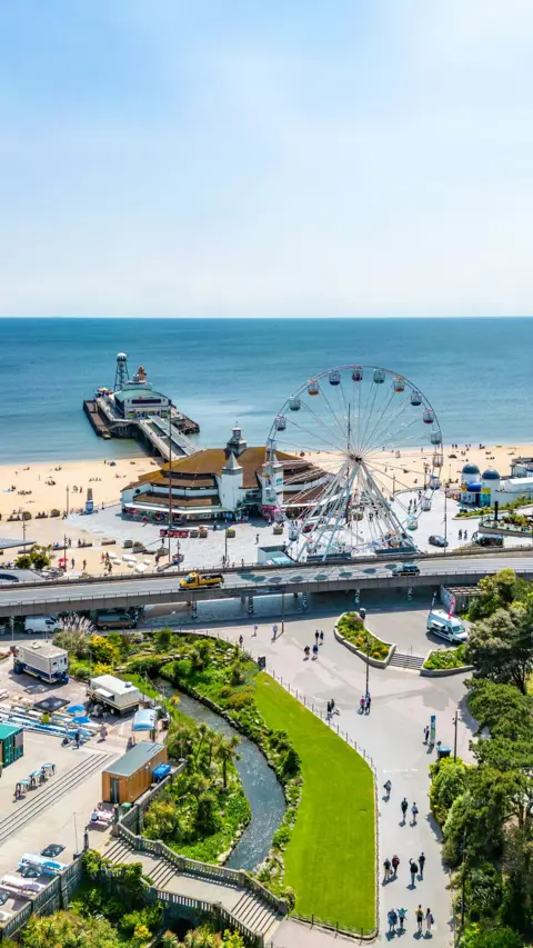 Bournemouth central gardens with the pier, beach and sea behind. The sky is clear blue and its sunny. People are walking along a path next to the grass and a stream. The photo has been taken from an elevated position.