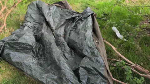 A discarded tarp of a tent is covering greenery. There is also litter left nearby the branch. 