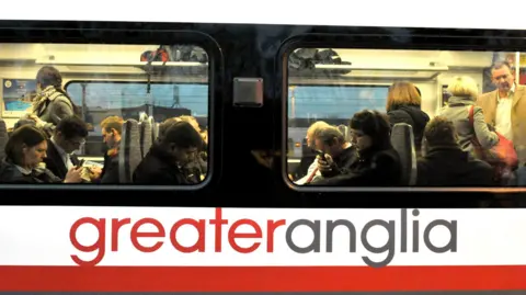 Commuters onboard a Greater Anglia carriage at Shenfield in Essex. There are men and women. Some are looking at their phone and others are standing.