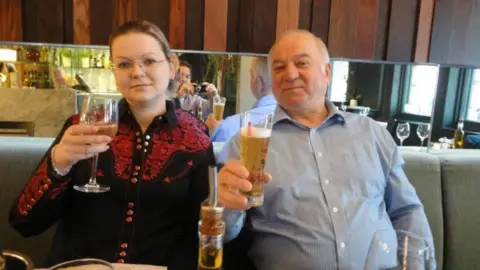 Sergey and Yulia Skripal sitting in a restaurant holding up their glasses full of alcohol. Yulia wears a black shirt with a red lace pattern and Sergey wears a blue shirt. 