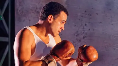 A black man in a boxer's stance, wearing a white vest and reddish brown gloves against a purple backdrop