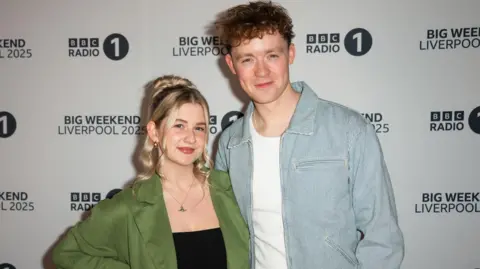Getty Images Sam MacGregor and Danni Diston at Radio 1 Big Weekend in May 2025. Danni (L) has her long blonde hair tied back and curled and wears a green blazer over a black top. Sam has short curly hair and wears a striped blue and white jacket over a white top.
