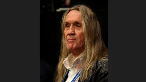 PA Media A head-and-shoulders picture of Nicko McBrain. He has long brown hair and is looking pensive. He is wearing an open-necked white shirt and a jacket.