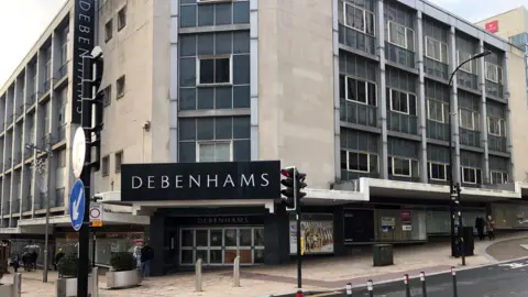 A large building, several storeys high, with a black sign and white letters saying Debenhams. The building is in a shopping precinct. It has empty windows and boarded up doors