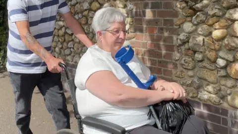 A woman, Pamela Bent, who has short grey hear, is wearing a white top and sitting in a wheelchair. She is holding a walking stick with a blue handle. A person wearing a blue striped top and grey jeans is pushing her in the wheelchair.