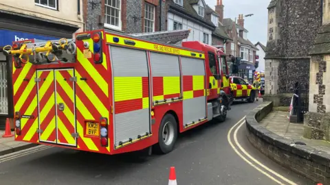 A fire engine is pictured parked in a narrow urban street, with cones behind it and a church on the right