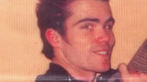 Martin Kelly smiling at the camera, he has dark hair and dark clothing on.