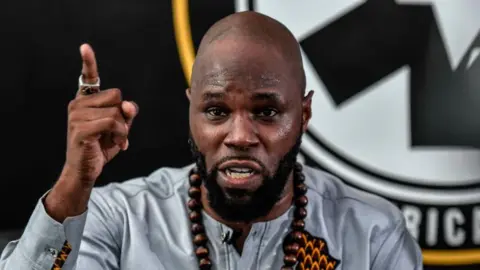 Kemi Seba is wearing a grey shirt, facing ahead. He is wearing a beaded wooden necklace and has his left arm raised in the air with his index finger pointing up. In the background is the partial logo of NGO Pan-Africanist Emergency