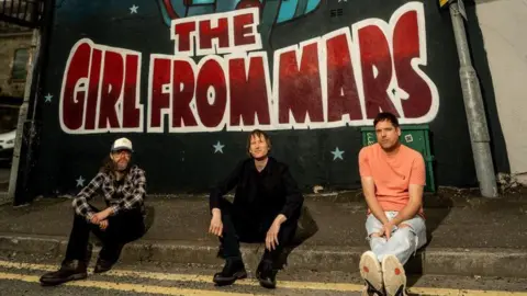 Roger Sargent Three men sitting casually on a pavement below a mural which says 'The Girl From Mars' in large red graffiti. They look relaxed.