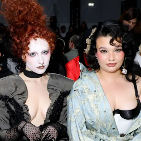 Getty Images Chappell Roan, wearing a low-cut grey jacket with black sheer gloves and white face make-up. Lola Young is wearing an off-the-shoulder light blue dress with a white and black corset underneath.