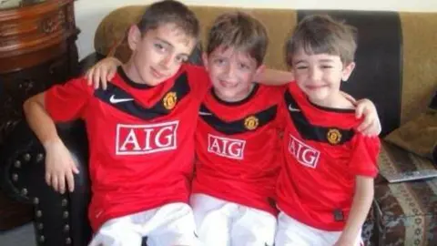 Bader Al-Akkad Three young boys, who are wearing Manchester United kits, have their arms around each other while they are sitting on a couch. 