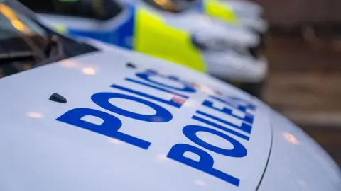 Generic image - extreme close up of a Police Scotland vehicle - white with blue text which reads "Police" and underneath "Poileas". In the background other police vehicles are visible but out of focus