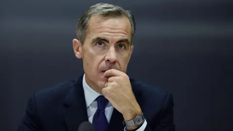 Getty Images Mark Carney, in a dark blue suit and tie and light shirt, listens during the Bank of England's Financial Stability Report press conference at the Bank of England