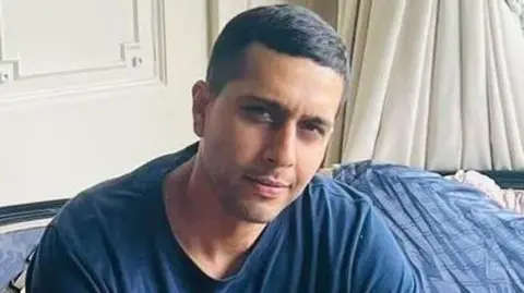 Kamran Aman, wearing a blue t-shirt, looks straight at the camera. He has short black hair. 