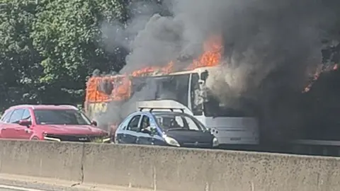 A white bus on fire. Two cars are driving in front of it.