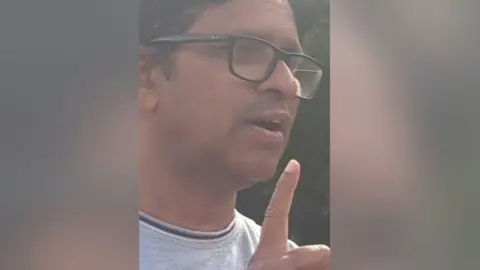 Close up of man's face he has dark, greying hair, is wearing glasses and is mid-speech and holding a finger in the air