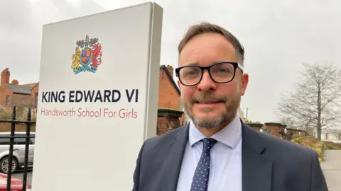 David Goodwin, standing outside the school where he is head teacher, next to a welcome sign.