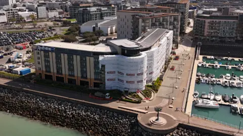BBC A picture of the front of the Radisson blu in Jersey. It is an all brick building with glass windows and plants in front of it. There is a large glass main entrance in the middle of the image. There is scaffolding surrounding the site.
