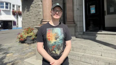 Filmmaker Lars standing in font of the Victor Hugo Centre. He is wearing a black shirt and cap. He is looking towards the camera. 