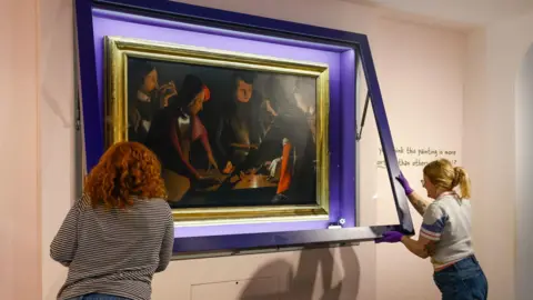 The Dice Players is a painting that features a close-up scene of five people rolling dice on a table. It is quite dark but there is a splash of colour on some of their clothing, including orange and purple. They are wearing formal and elegant clothing. It is inside a large gold frame, which is being placed back into its case on a gallery wall at Preston Park Museum by two members of staff. The two women, pictured from behind, are closing the case.