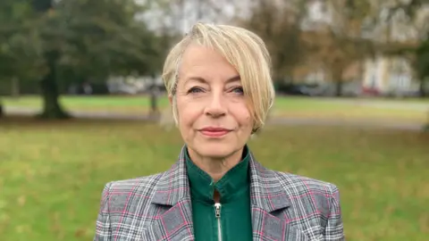 A woman with short blonde hair in the park. She wears a green zip-up top with the collar up and a grey checked jacket on top. 