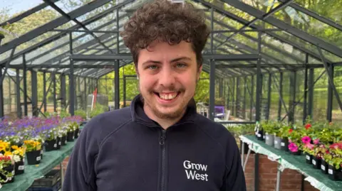 Ethan smiling into the camera while standing in one of the greenhouses. He has brown, curly hair and is wearing a blue zip-up fleece which reads 'Grow West' on the right of his chest. There are tables on either side of him lined with pots with different coloured flowers in them. On the right they are pink and on the left they are yellow, purple and pink.