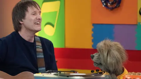 Tim and the CBeebies dog Dodge sitting together singing. They're sitting in a multi-coloured room and Tim is holding a guitar. He's wearing a navy top.