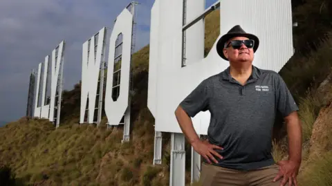 Getty Images A man in a trilby hat and sunglasses poses in front of the Hollywood Sign. He has one hand on his hip, the other resting on his opposite leg. The off-camera flash and wide angle creates a dynamic contrast between the subject and the structure in the background.