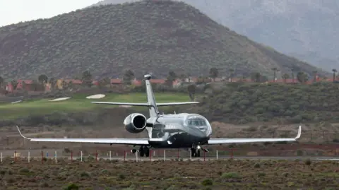 A dark coloured plane taking off on a runway with a green hill in the background. The photo is from the front looking towards the executive-style two-engine aircraft.