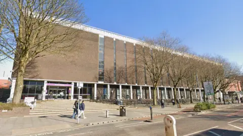 Google A long university building in front of a tree-lined street with people walking along the road on a clear day.